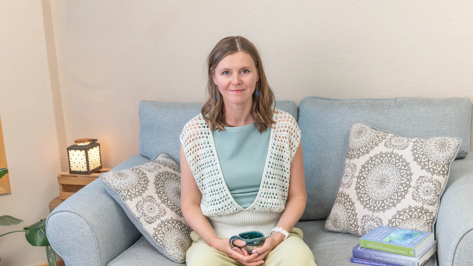Portrait of Anna seated on a sofa in her therapy room, holding a ceramic mug